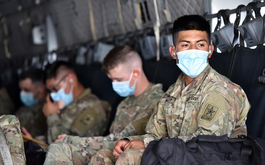 A soldier looks out of the loading ramp of a C-27 aircraft while he and his fellow soldiers wear face masks and maintain social distance.