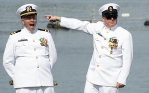 Two men in dress white uniforms stand near water.