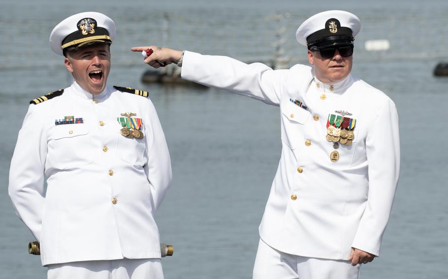 Two men in dress white uniforms stand near water.