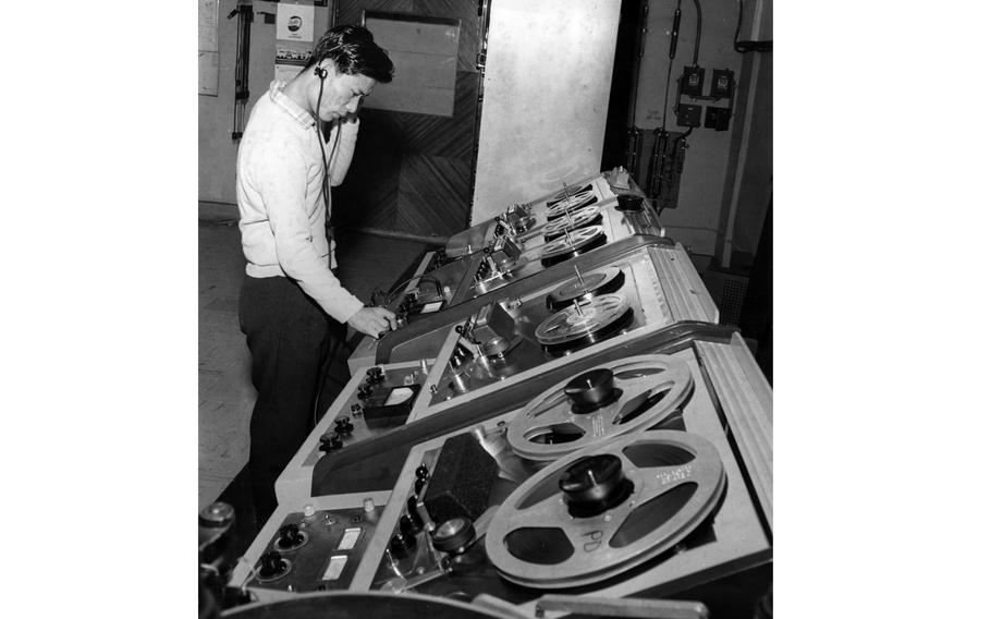 A black-and-white, 1960s-era image of a Japanese man wearing headphones and standing over a large radio control panel.