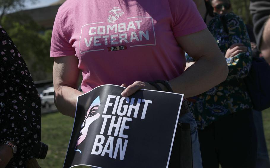 A close-up view of a person’s torso wearing a pink shirt that reads “Combat Veteran” while holding a sign at the waist that reads “Fight  The Ban.”