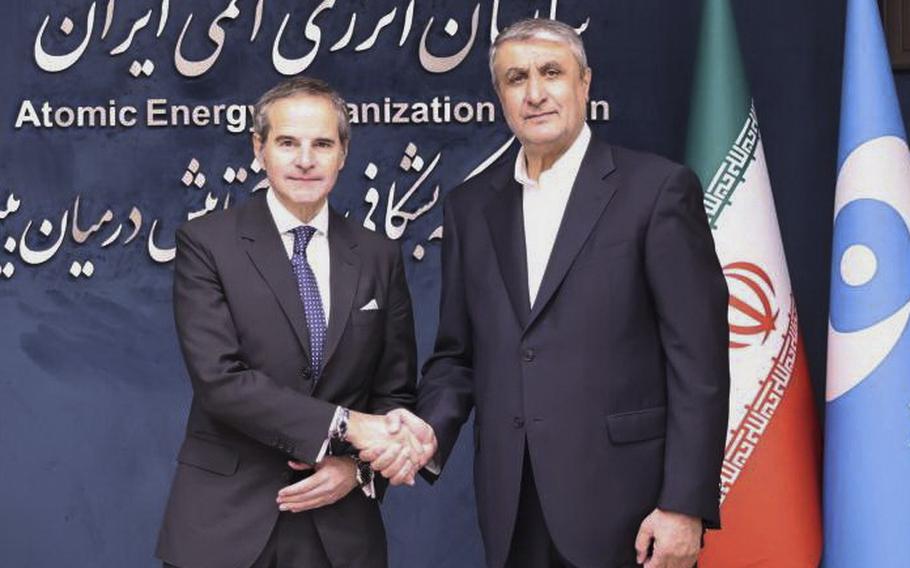 Two men in dark suits shake hands while posing next to an Iranian flag.