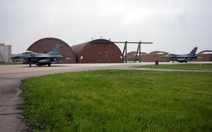 Two F-16 fighter jets are positioned on a concrete runway in front of brown curved aircraft hangars at a military air base, with green grass visible in the foreground and an overcast sky above. 