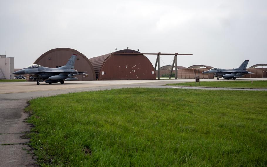 Two F-16 fighter jets are positioned on a concrete runway in front of brown curved aircraft hangars at a military air base, with green grass visible in the foreground and an overcast sky above. 