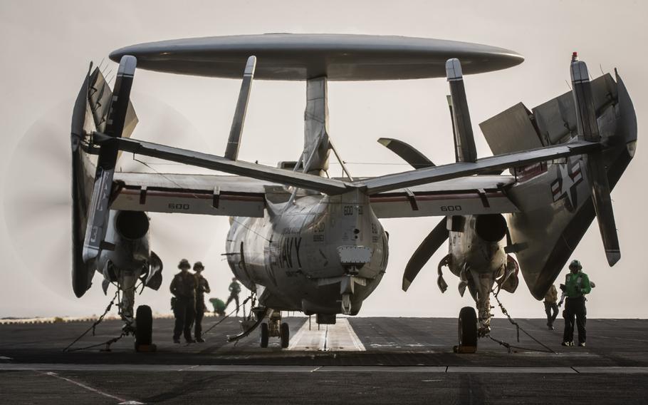 An aircraft sits on the deck of a ship, while two crew members stand behind it.