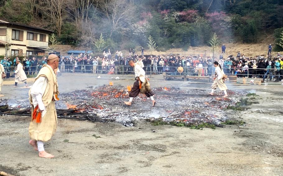 Japanese monks in traditional attire walk across a patch of burning, smoldering coals while a crowd of spectators watches.