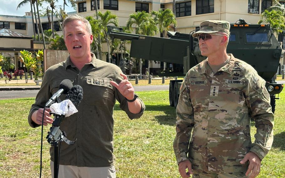 A man in an olive-green button-down shirt speaks into a microphone while standing in a grassy area next to an Army officer in camouflage uniform and in front of an armored vehicle.
