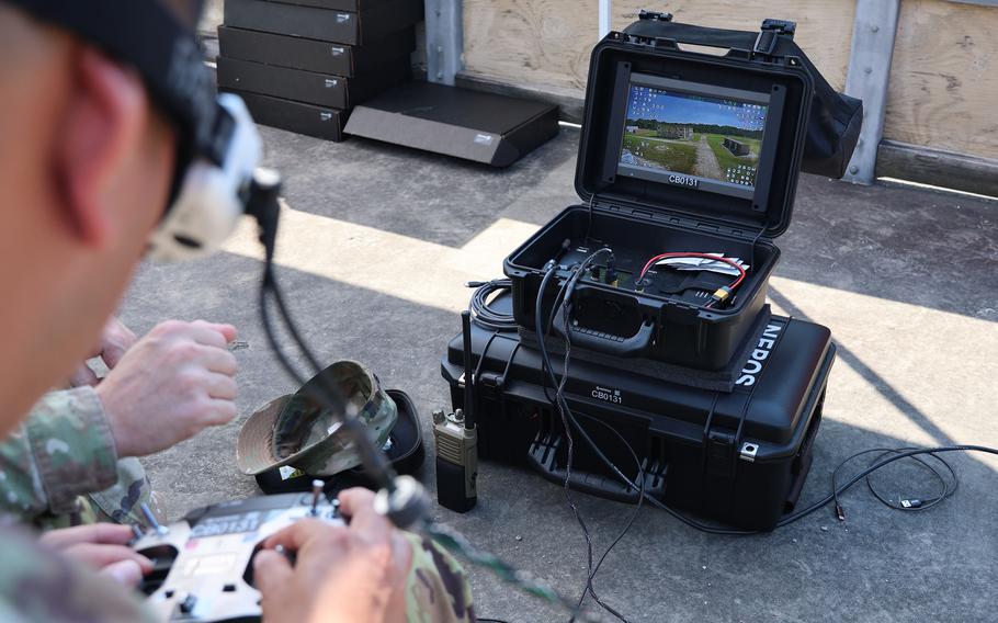 A soldier operates a remote control while wearing a VR headset; both are plugged into a larger machine in front of him.