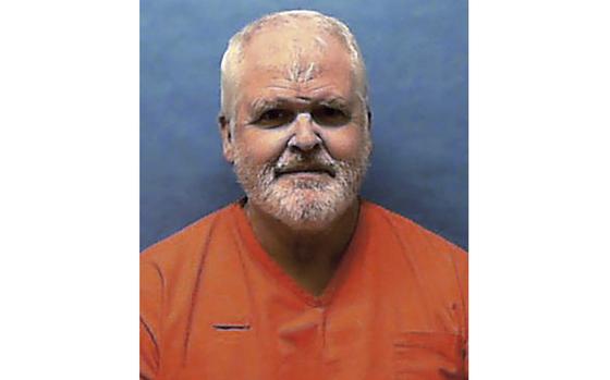 A man in an orange prison jumpsuit poses in front of a blue wall.