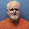 A man in an orange prison jumpsuit poses in front of a blue wall.