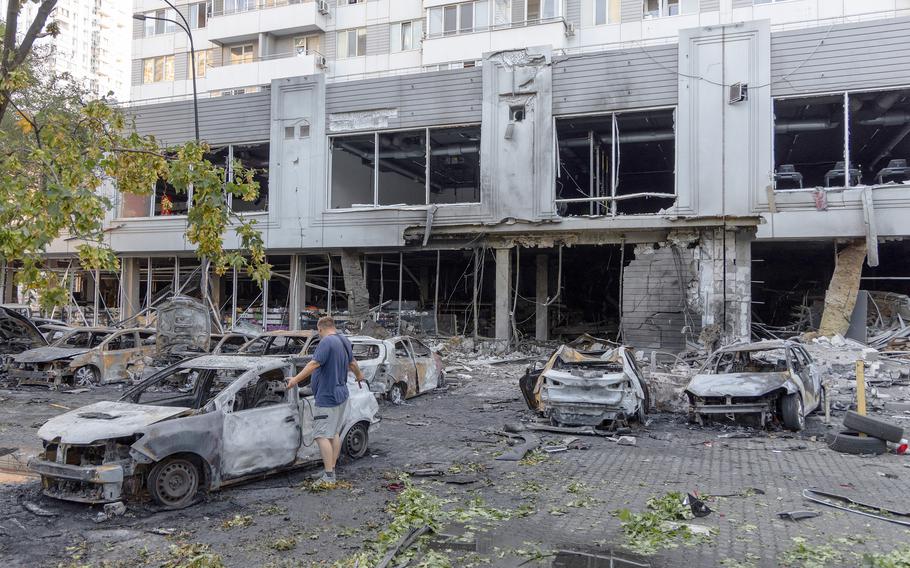 A man looks at a burnt car in a lot with other burnt and damaged cars.