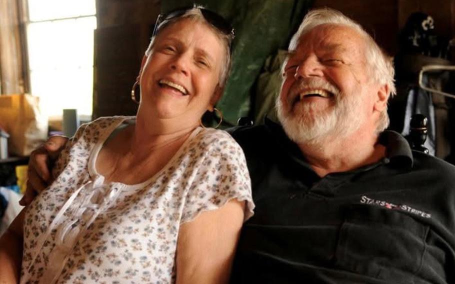 An elderly couple, seen from the waist up, laugh while seated next to each other.