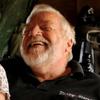 An elderly couple, seen from the waist up, laugh while seated next to each other.