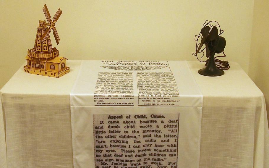 A table covered by a white cloth with a strip of cloth carrying newspaper print on the table’s center, a model windmill on the left and a fan on the right.