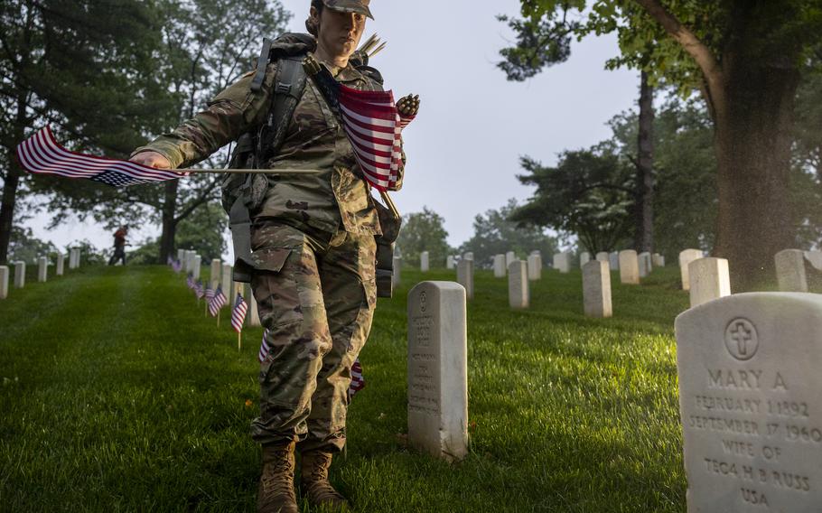 A soldier with multiple flags prepares to place one in front of a headstone.