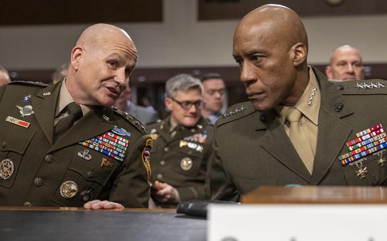 Two military officers in dress uniforms lean towards each other while seated at a table in a congressional hearing room.