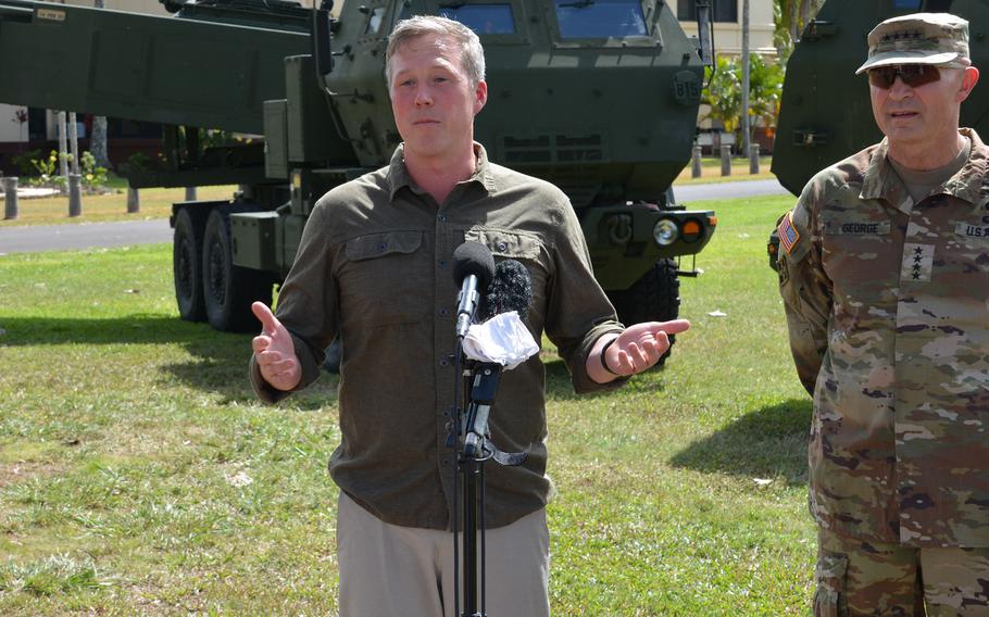 A man in an olive-green button-down shirt speaks into a microphone while standing in a grassy area next to an Army officer in camouflage uniform and in front of an armored vehicle.