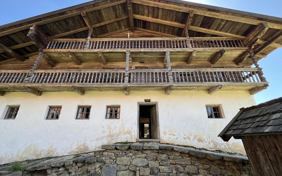 A traditional alpine farmhouse with white plastered walls, small barred windows, and a wooden upper floor with a long balcony made of carved wooden railings, set on a stone foundation under a wide gabled roof.