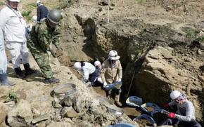 People wearing hardhats and gloves dig and sift through soil as part of an effort to recover World War II remains. The area around is partially excavated, with earth and rocks exposed.