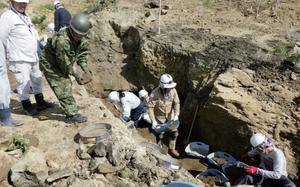 People wearing hardhats and gloves dig and sift through soil as part of an effort to recover World War II remains. The area around is partially excavated, with earth and rocks exposed.