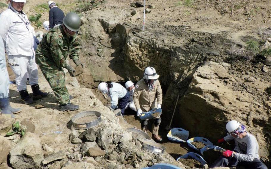 People wearing hardhats and gloves dig and sift through soil as part of an effort to recover World War II remains. The area around is partially excavated, with earth and rocks exposed.