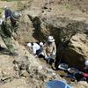 People wearing hardhats and gloves dig and sift through soil as part of an effort to recover World War II remains. The area around is partially excavated, with earth and rocks exposed.