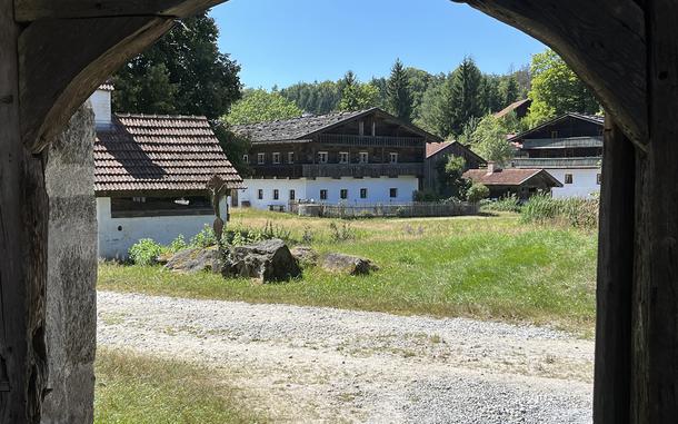 View through a stone archway of a traditional Alpine village with white buildings featuring dark wooden balconies and red tile roofs, set against forested hills under a clear blue sky.