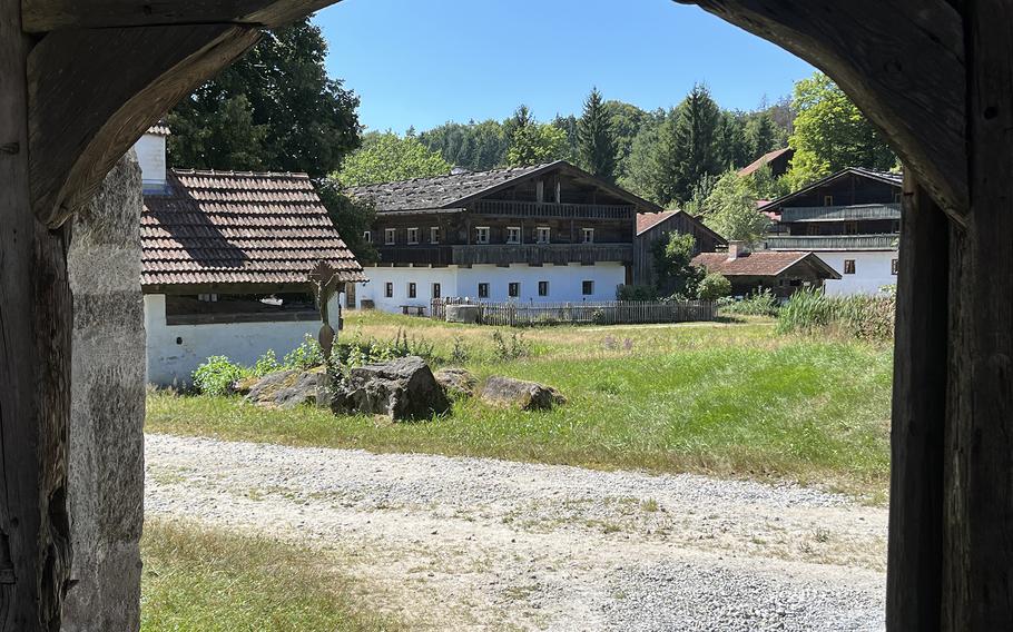 View through a stone archway of a traditional Alpine village with white buildings featuring dark wooden balconies and red tile roofs, set against forested hills under a clear blue sky.