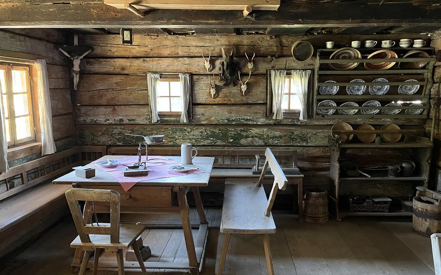 Interior of a traditional Alpine kitchen with log walls, wooden furniture, shelving displaying pottery and dishes, hunting trophies mounted on walls, small windows with curtains, and rustic wooden table and chairs.