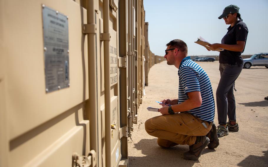 Two people inspect large containers, one kneeling and the other standing.