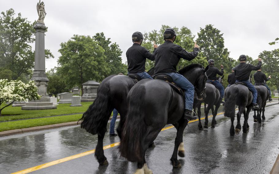 Soldiers ride side by side in a line, hands up, as they pass a statue.