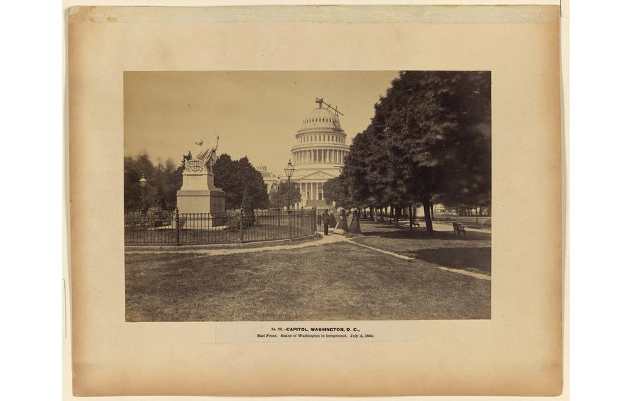 A view from behind of a statue of George Washington by Horatio Greenough in front of the U.S. Capitol under construction on July 11, 1863. A group of sightseers stands nearby. 