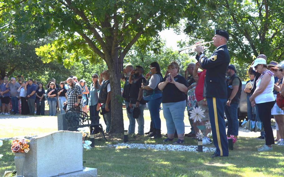 A soldier plays a bugle as people are gathered under a tree near a headstone.