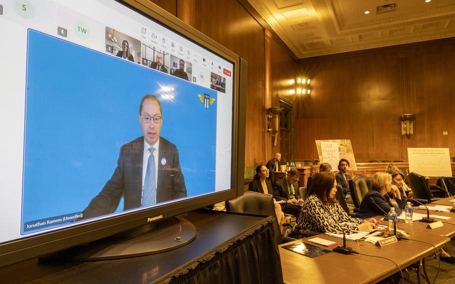 Large TV screen shows a man speaking and people seated next to the screen listening. 