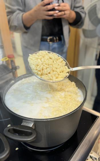 Spaetzle noodles in metal spoons above a large black pot of boiling water and noodles