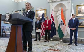 A Japanese government official stands at a podium in an ornate room and speaks into a microphone, while Indian, Australian and U.S. officials stand in a line behind him.