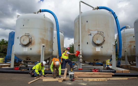 Workers in orange and yellow reflective vests crouch down and stand next to two large water tanks.