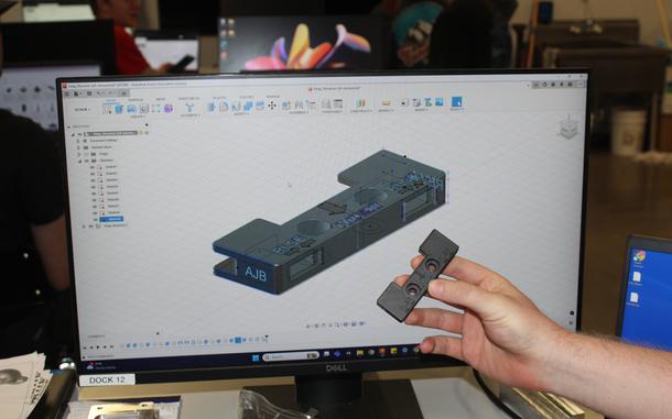 A hand holds a gray object in front of a computer screen displaying a representation of that object.