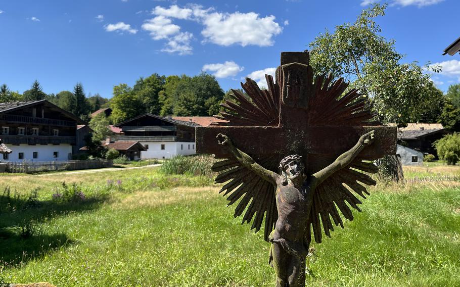 A rustic metal crucifix depicting Jesus on the cross stands in the foreground of a scenic rural village. Behind it are traditional alpine-style wooden houses with white walls and sloped roofs, surrounded by lush green meadows, trees, and a bright blue sky with scattered clouds.