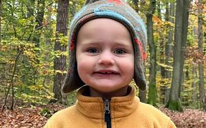 Caleb Anders, a toddler in a yellow jacket and dinosaur hat, smiles as he poses for a photo in the woods.