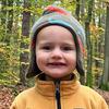 Caleb Anders, a toddler in a yellow jacket and dinosaur hat, smiles as he poses for a photo in the woods.