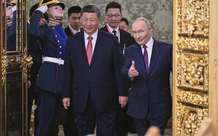Vladimir Putin walks next to Xi Jinping as aids follow behind them, with Putin gesturing a hand forward.
