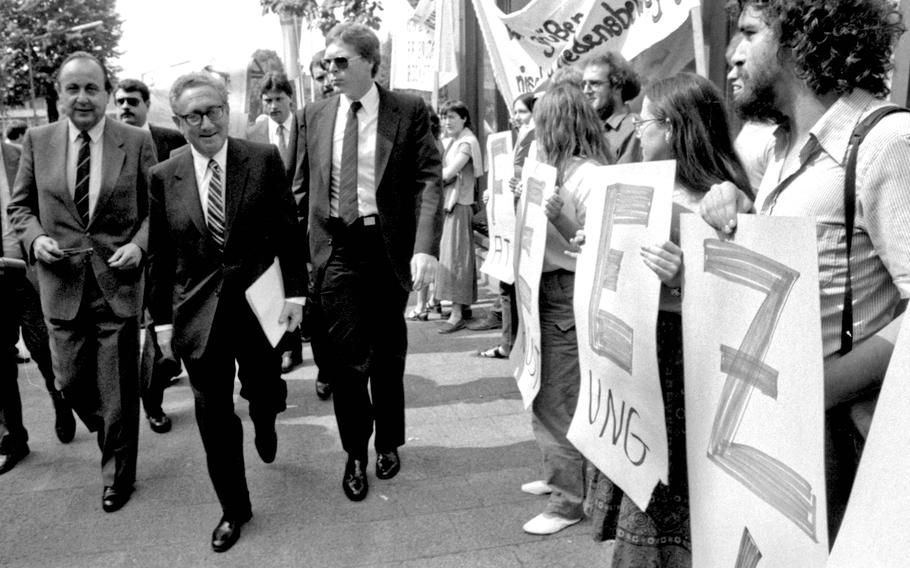Former Secretary of State Henry Kissinger walks past a line of protesters in Worms, Germany, on his way to speak at a German-American tricentennial ceremony on June 12, 1983. At left is German Foreign Minister Hans-Dietrich Genscher. The protesters are holding letters forming the word “freeze,” in connection with nuclear disarmament.