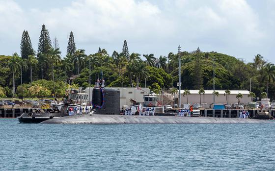 The top of a submarine is seen above the water line next to a pier with a tropical treeline in the background.