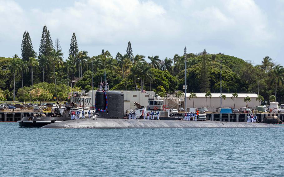 The top of a submarine is seen above the water line next to a pier with a tropical treeline in the background.