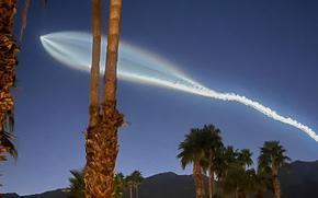 A trail of light and smoke cuts through a dark blue sky, which is also partially obscured by palm trees.