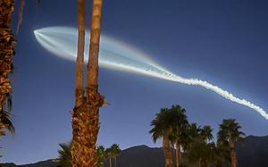 A trail of light and smoke cuts through a dark blue sky, which is also partially obscured by palm trees.