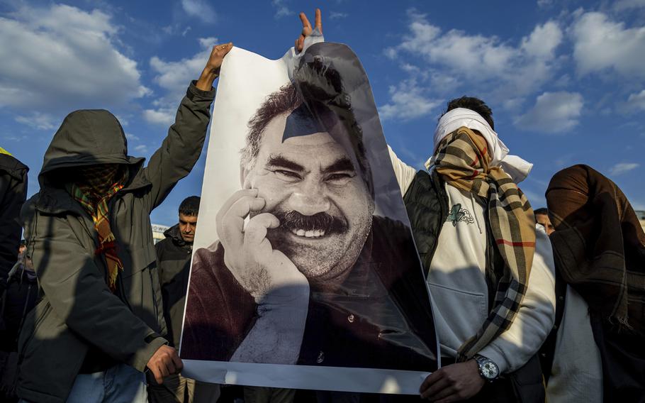 Men with their faces covered by hoods and scarves hold a large poster of a man’s face.