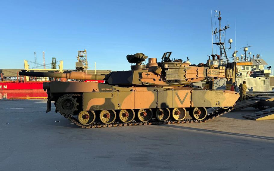 A camouflage painted tank sits on a concrete pier next to a ramp leading onto a cargo vessel.
