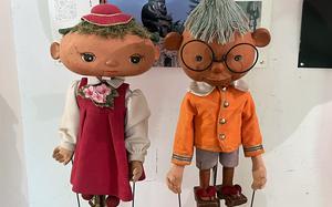 Two puppets, one with a red dress and hat and the other with an orange shirt, sit on a display in front of a white wall.